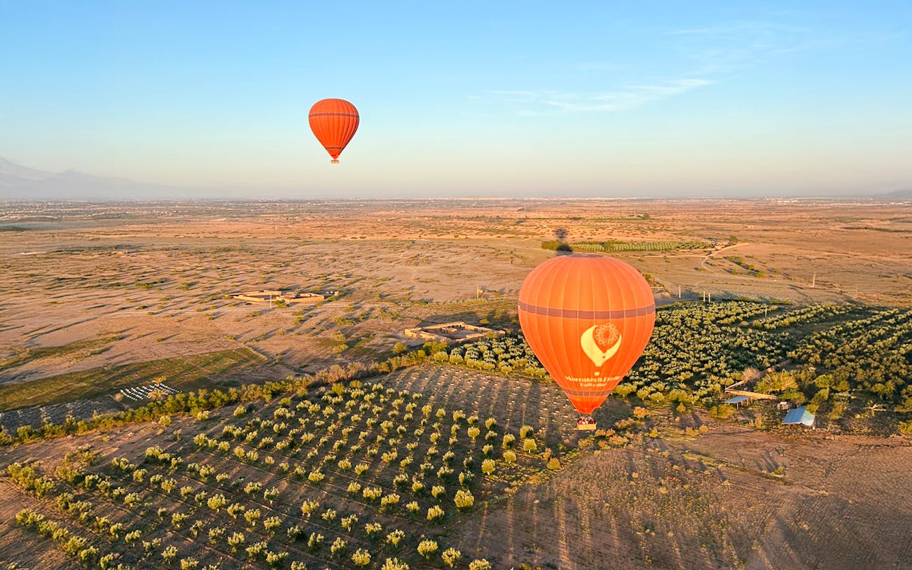 Hot air balloons floating over Marrakech landscape with fields and distant mountains.