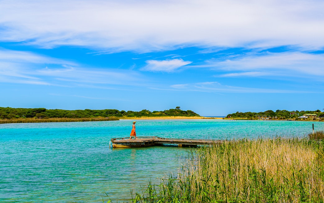 Woman walking on Anglesea River boardwalk with scenic view of water and greenery.