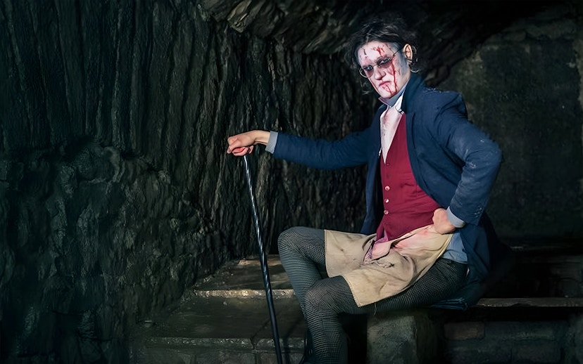 Victorian-era actor in makeup and costume in a dimly lit underground setting for paranormal tour.