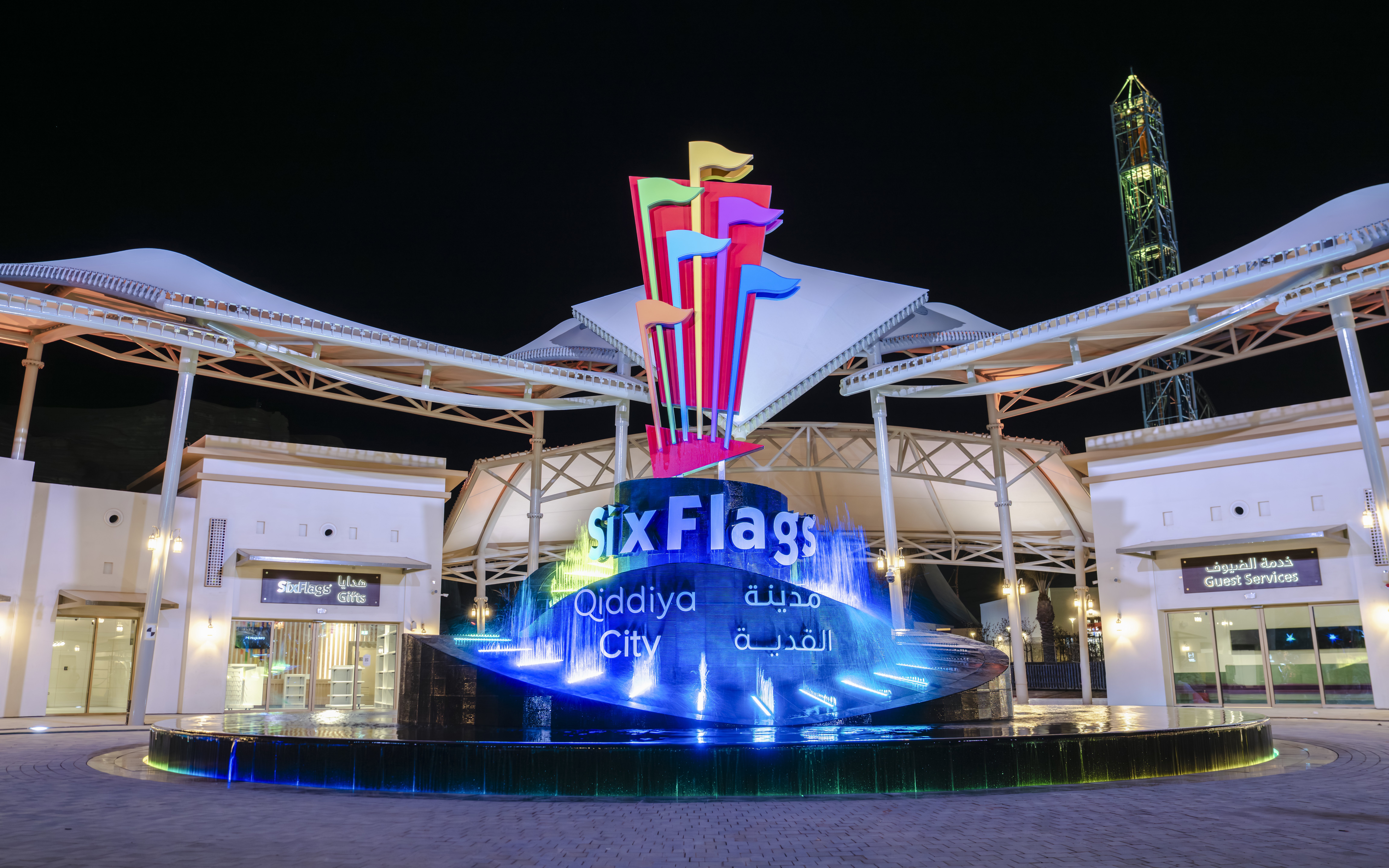 Six Flags Qiddiya City entrance with illuminated sign and colorful flags at night.