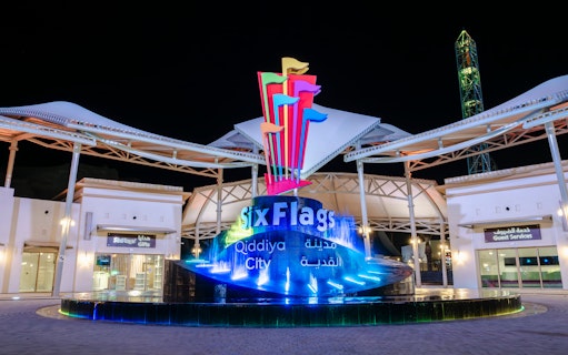 Six Flags Qiddiya City entrance with illuminated sign and colorful flags at night.