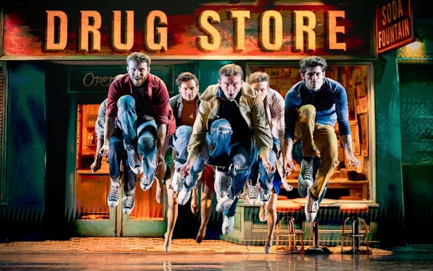 Performers leaping in front of a vintage drugstore sign during a dance show.