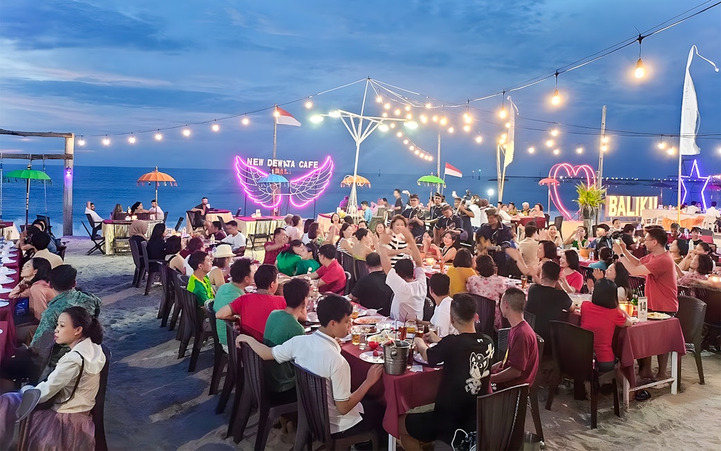 Crowded beachfront dining at New Dewata Cafe, Jimbaran Bali, with seafood dishes and ocean view.