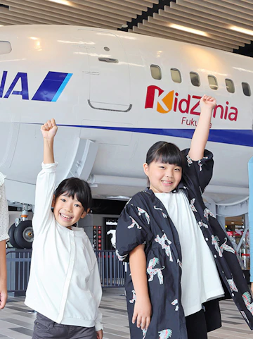 Children raising arms in front of KidZania Fukuoka airplane exhibit in Tokyo.