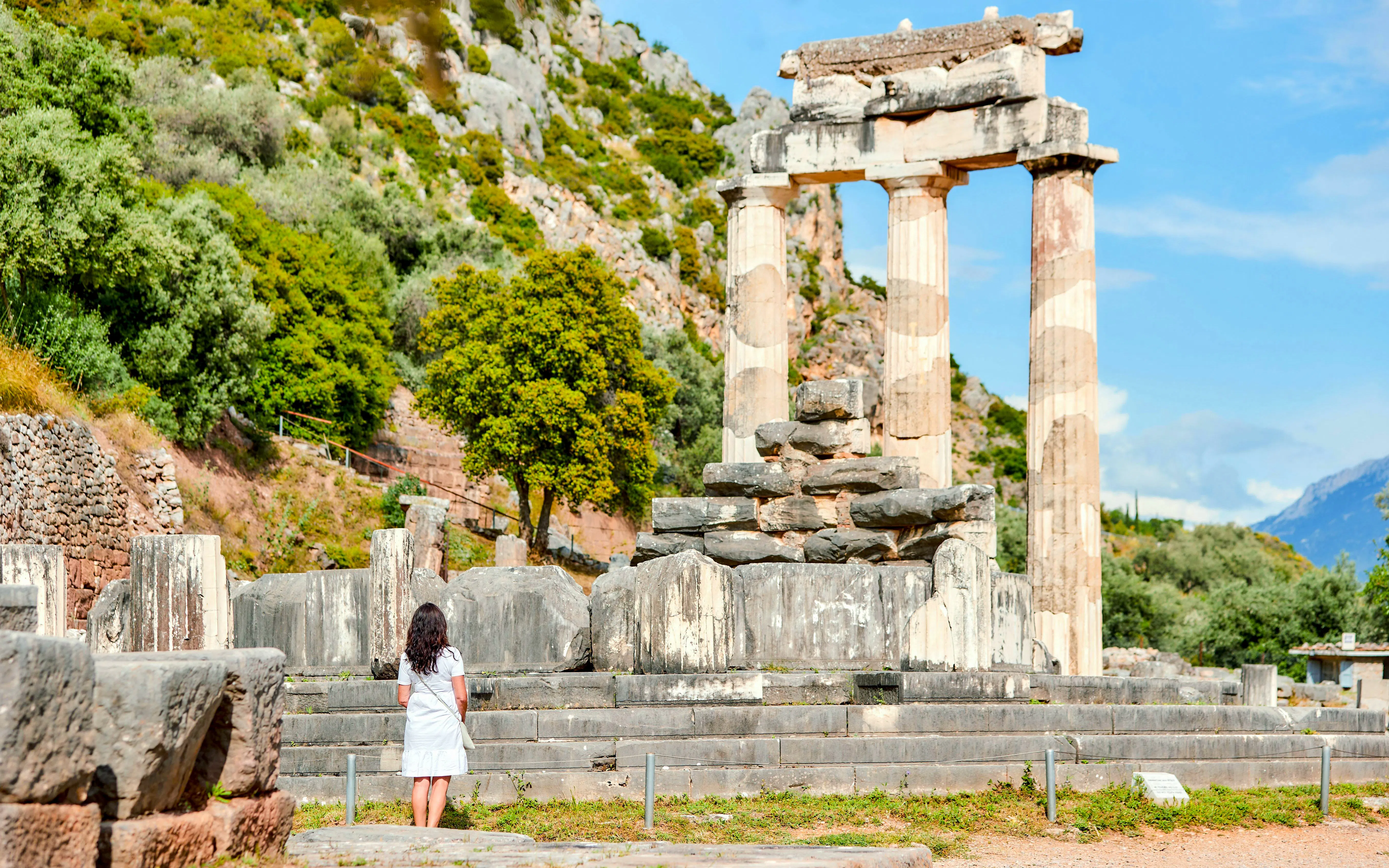 Woman in white dress at Athena Pronaia temple, Delphi, Greece, sunny day, UNESCO site.