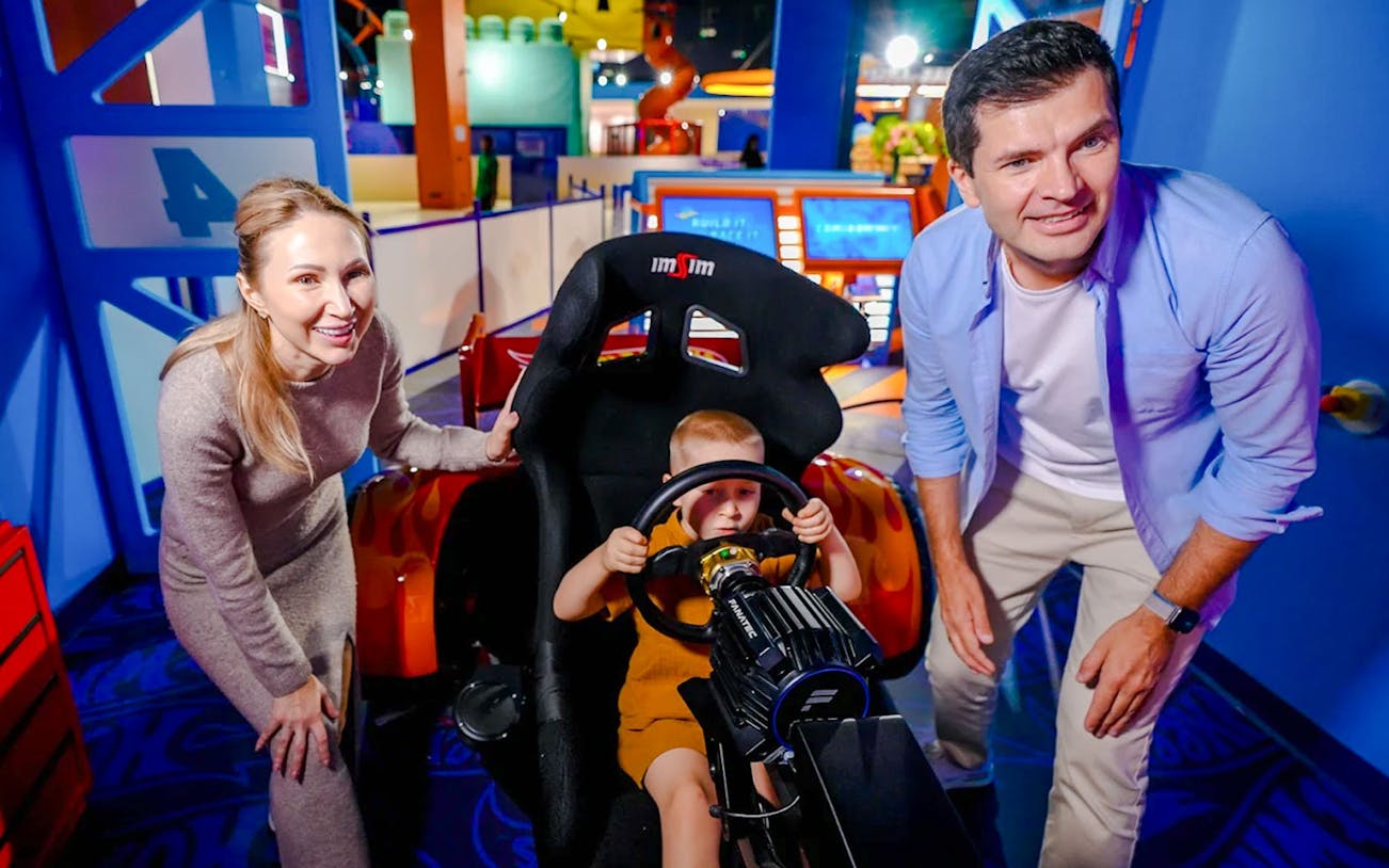 Child playing in a racing simulator at Mission: Play! by Mattel, with adults watching.