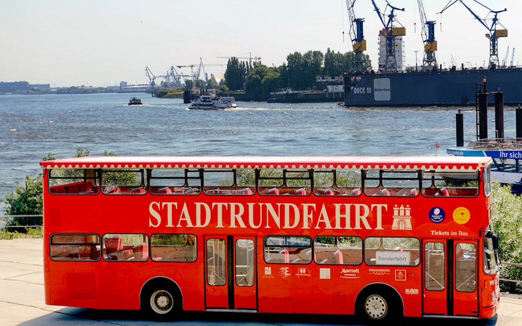 Red double-decker tour bus by the river in Hamburg, Germany with cranes in the background.