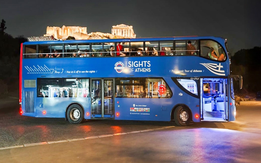 Open double-decker bus in Athens with Acropolis view at night.