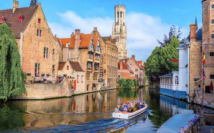 Boat tour on Bruges canal with Belfry of Bruges in background, Belgium.