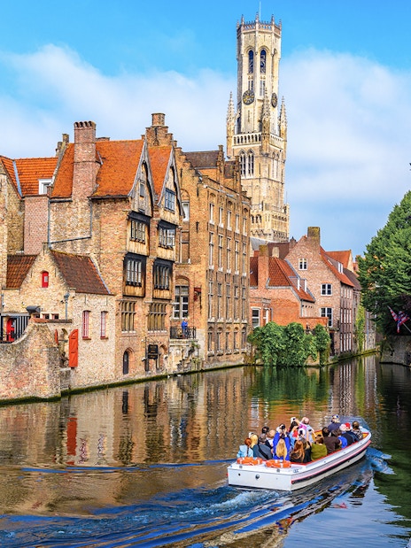 Boat tour on Bruges canal with Belfry of Bruges in background, Belgium.