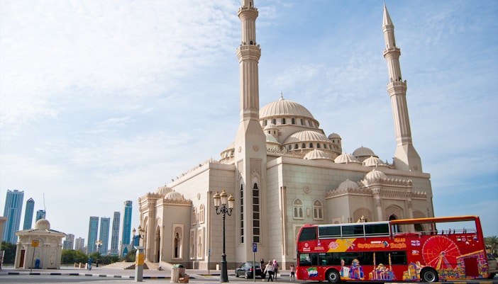 Sharjah mosque with a red Hop-On Hop-Off bus in front, city skyline in the background.