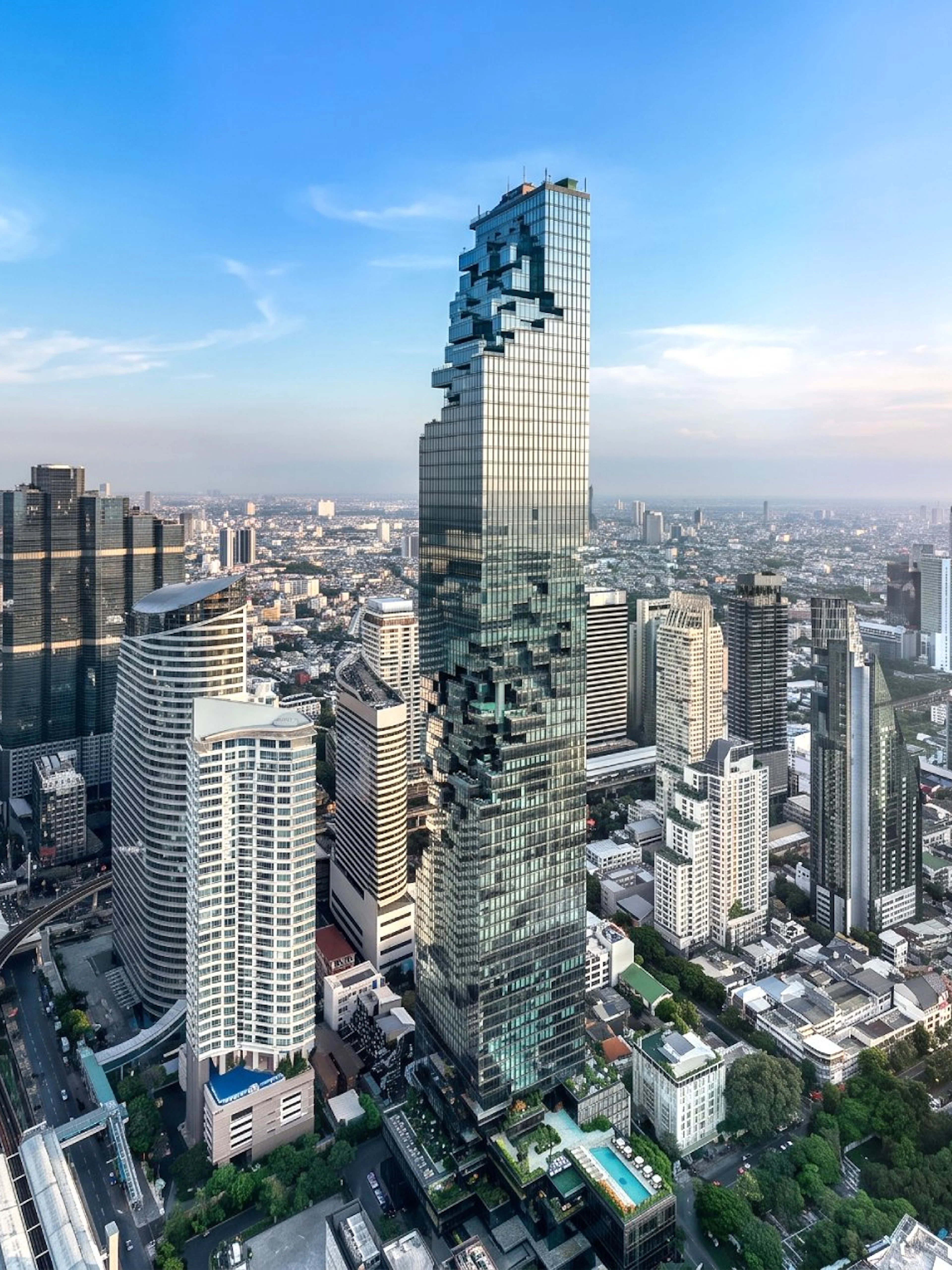 High-angle view of King Power Mahanakhon skyscraper in Bangkok cityscape under clear blue sky.