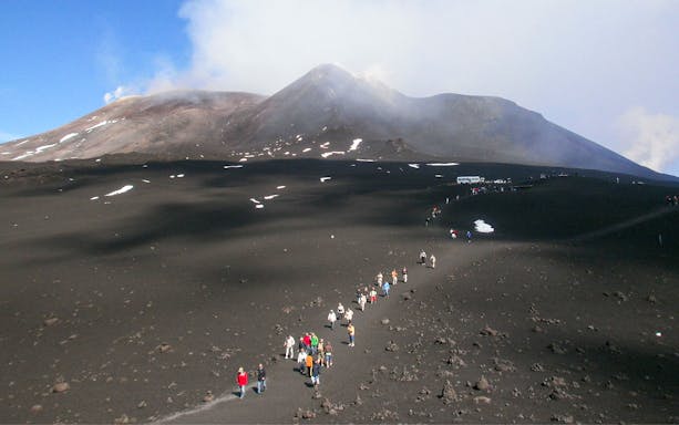 Group hiking on volcanic terrain of Mount Etna, Sicily, with smoke in the background.
