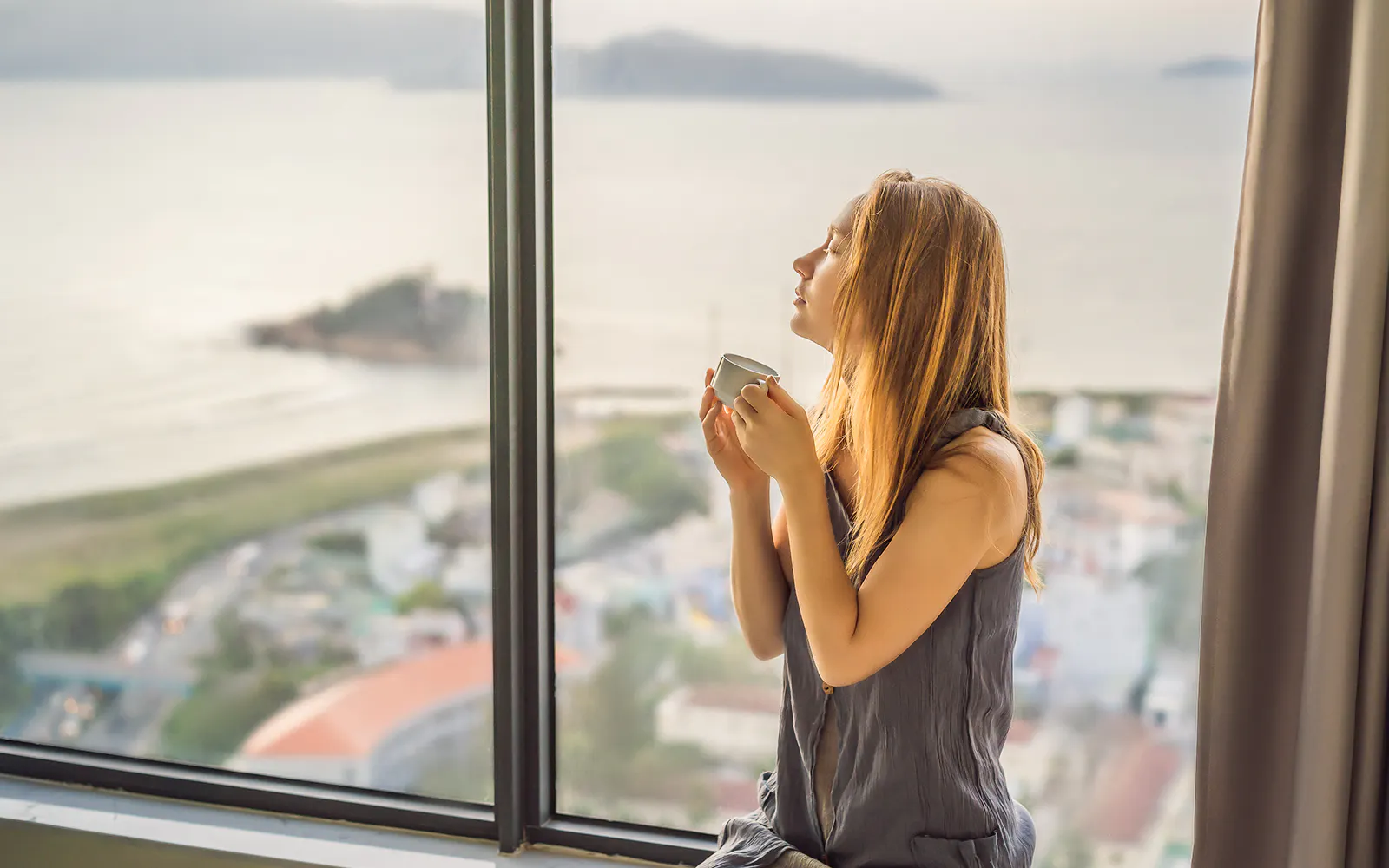 Young woman is drinking coffee in the morning on the background of a window with a sea view