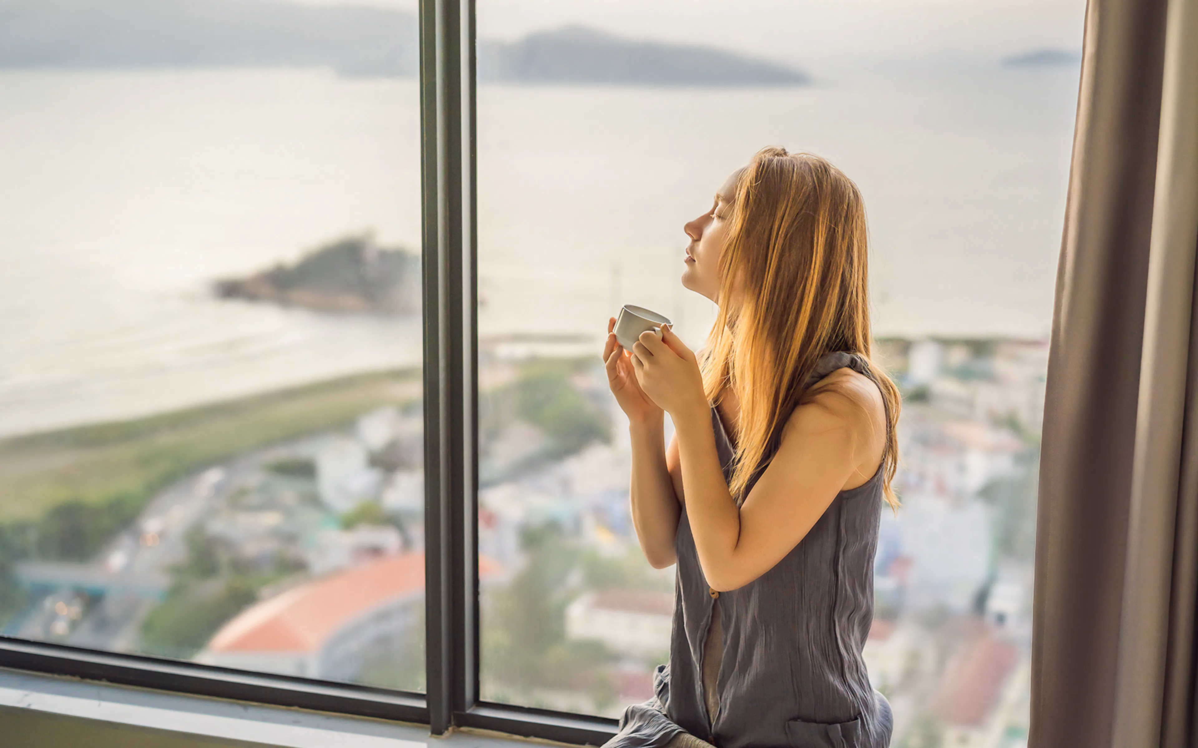 Young woman is drinking coffee in the morning on the background of a window with a sea view