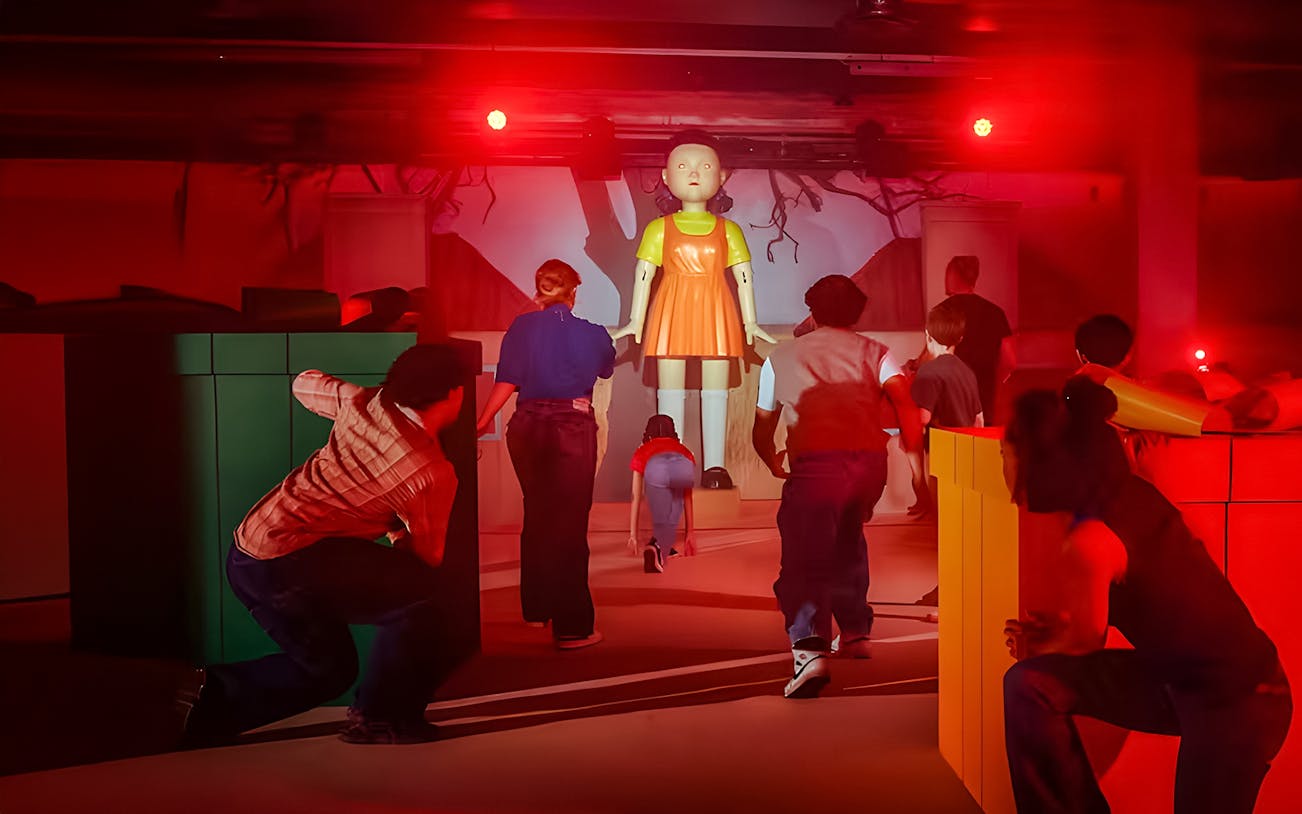 Participants in a Squid Game experience in London, crouching near a giant doll.