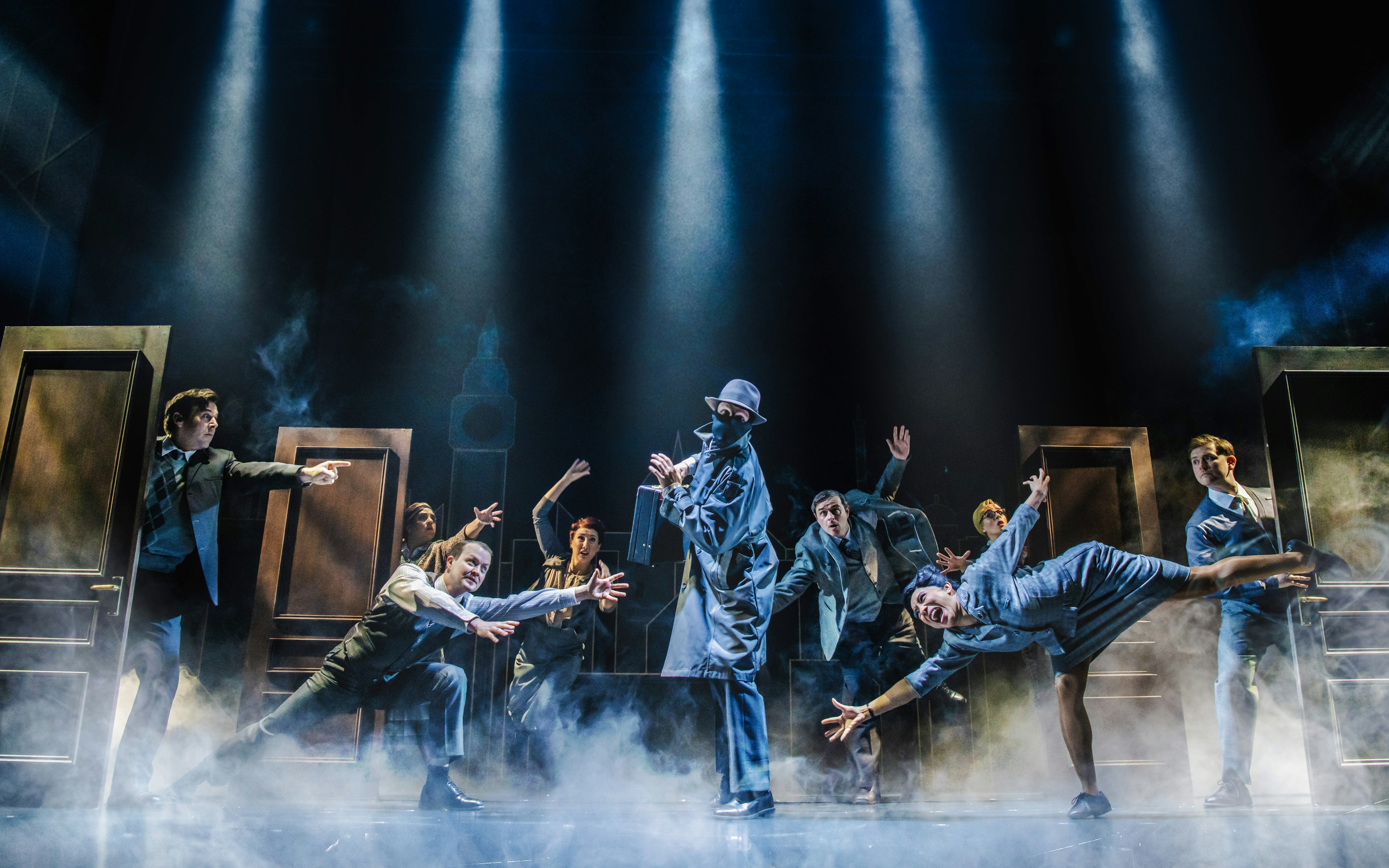 Actors performing a comedic spy scene on stage with dramatic lighting.