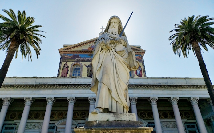 Colossal statue of Saint Paul at the Papal Basilica, Rome, flanked by palm trees.