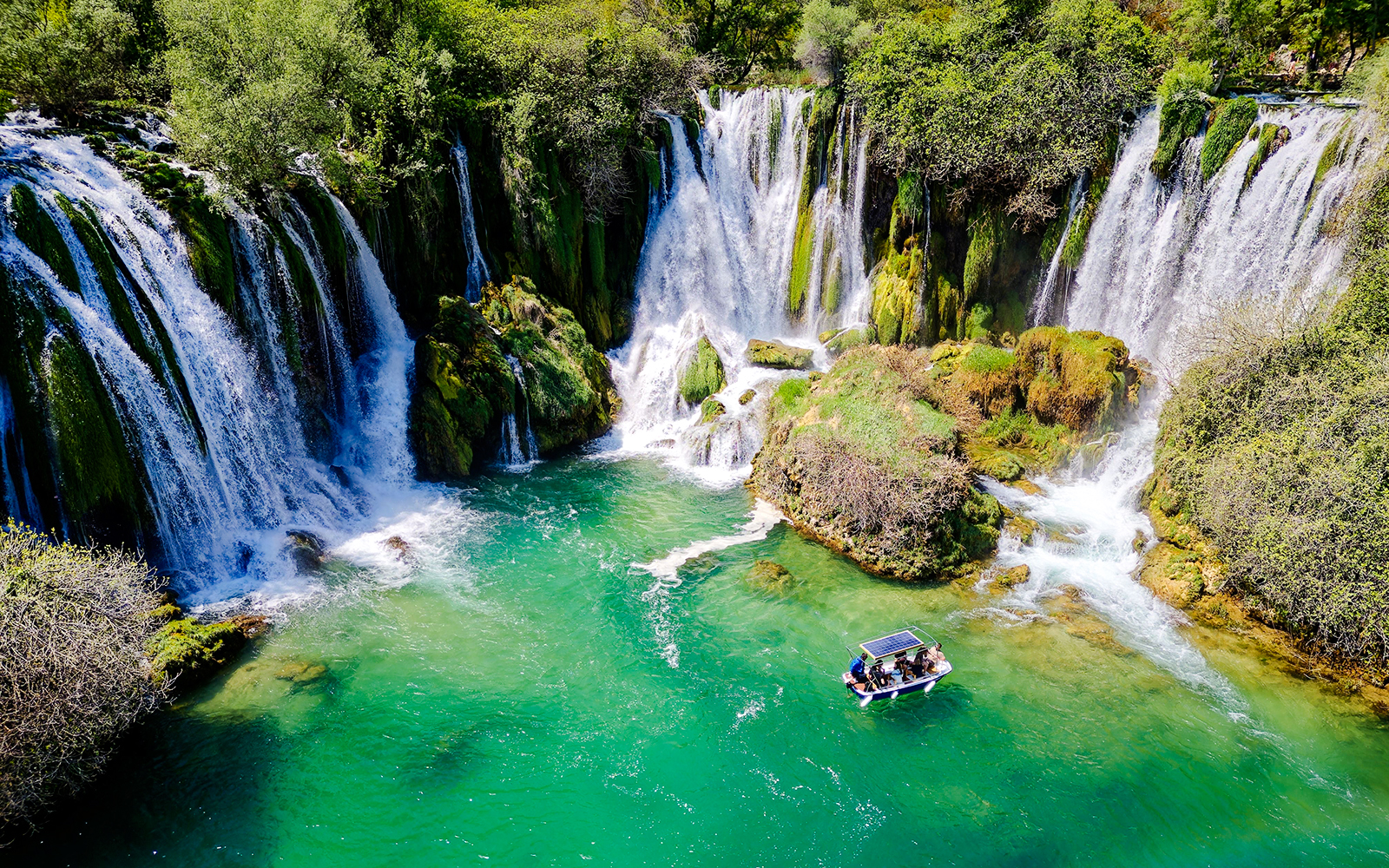 Aerial view of Kravice Waterfall with a boat on turquoise water.