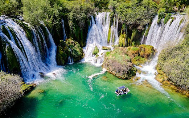 Aerial view of Kravice Waterfall with a boat on turquoise water.