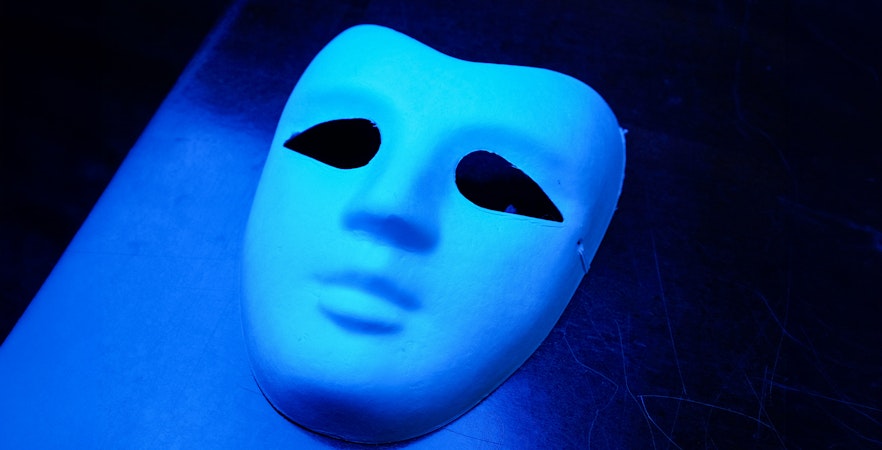 Blue theatrical mask with black eye holes on a dark surface.