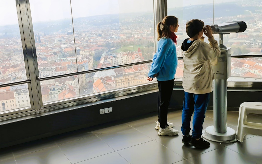 Children using a telescope inside Zizkov TV Tower with Prague city view.