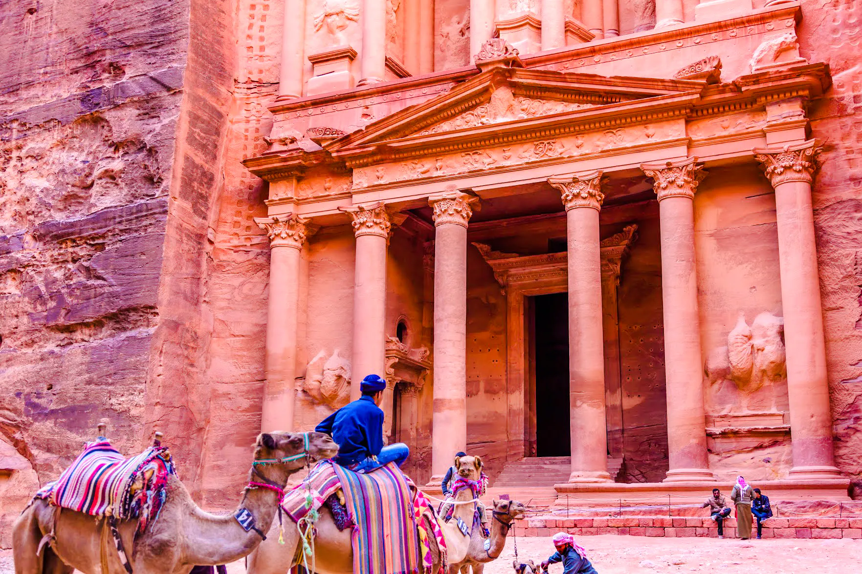 Camels in front of the Treasury at Petra, Jordan, with tourists exploring the ancient site.