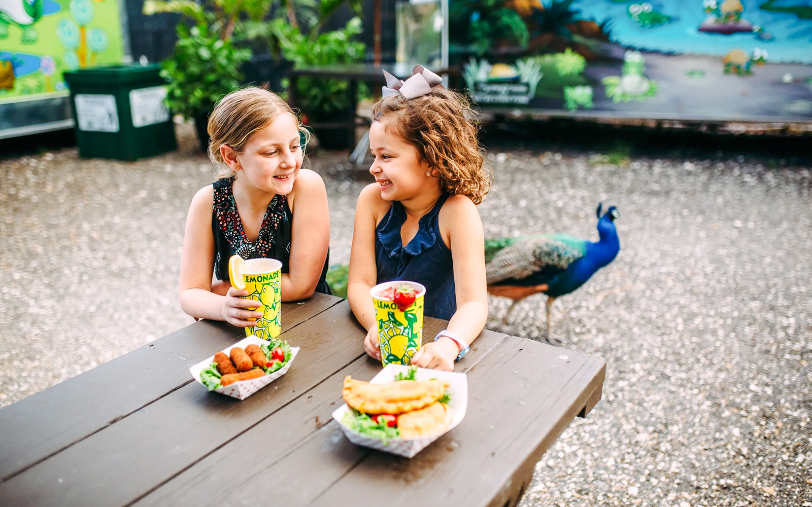 Kids enjoying a meal at Sawgrass Recreation Park with a peacock in the background.