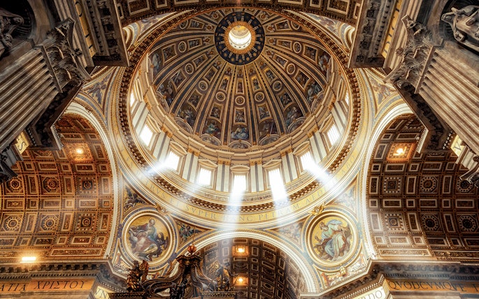 Dome and ornate ceiling of St. Peter’s Basilica with sunlight streaming through windows.