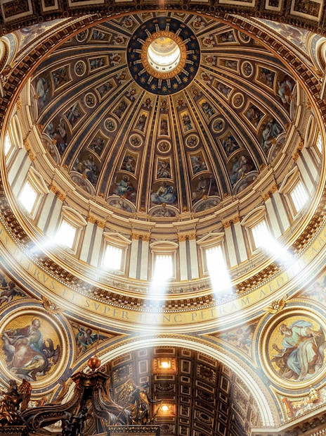 Dome and ornate ceiling of St. Peter’s Basilica with sunlight streaming through windows.