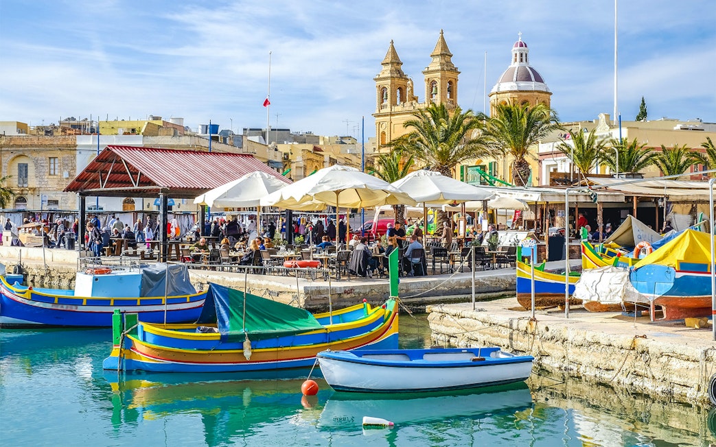 Colorful boats in a Maltese harbor with a church and outdoor market in the background.