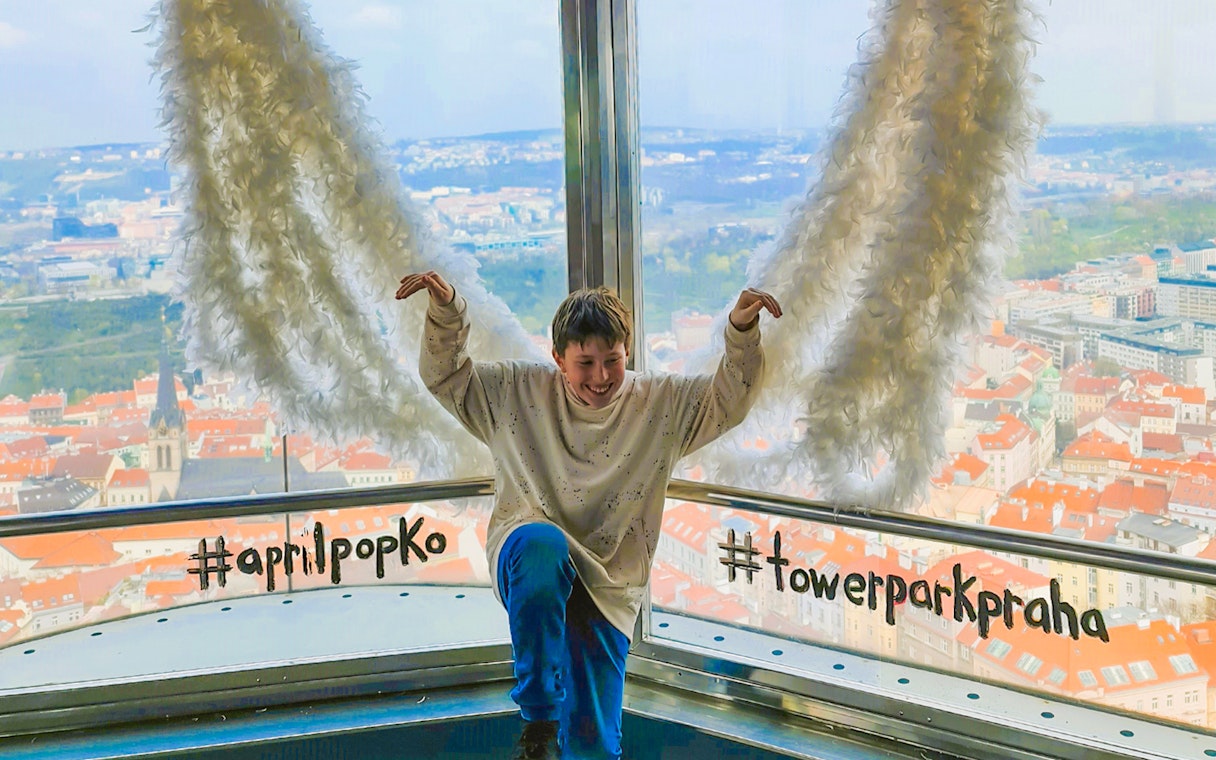 Person posing with feather wings at Žižkov Television Tower, Prague, overlooking cityscape.