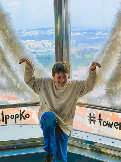 Person posing with feather wings at Žižkov Television Tower, Prague, overlooking cityscape.