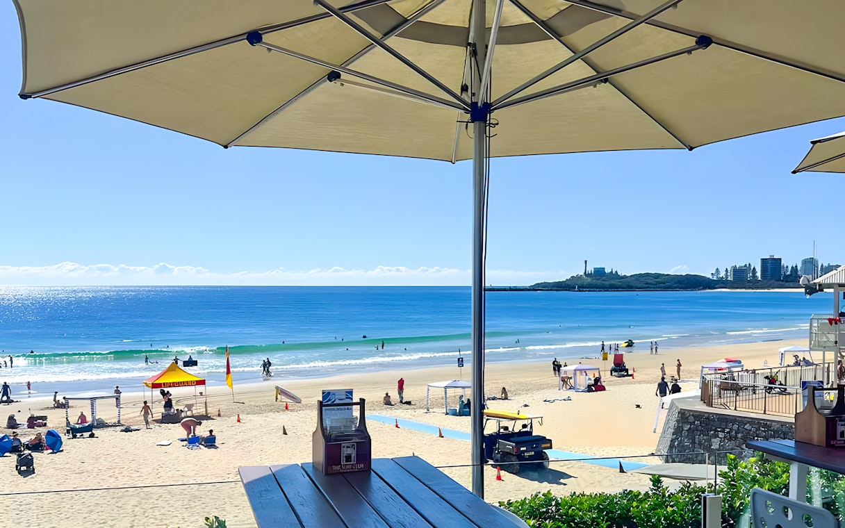 Mooloolaba Beach view with umbrellas, lifeguard station, and people enjoying the ocean.