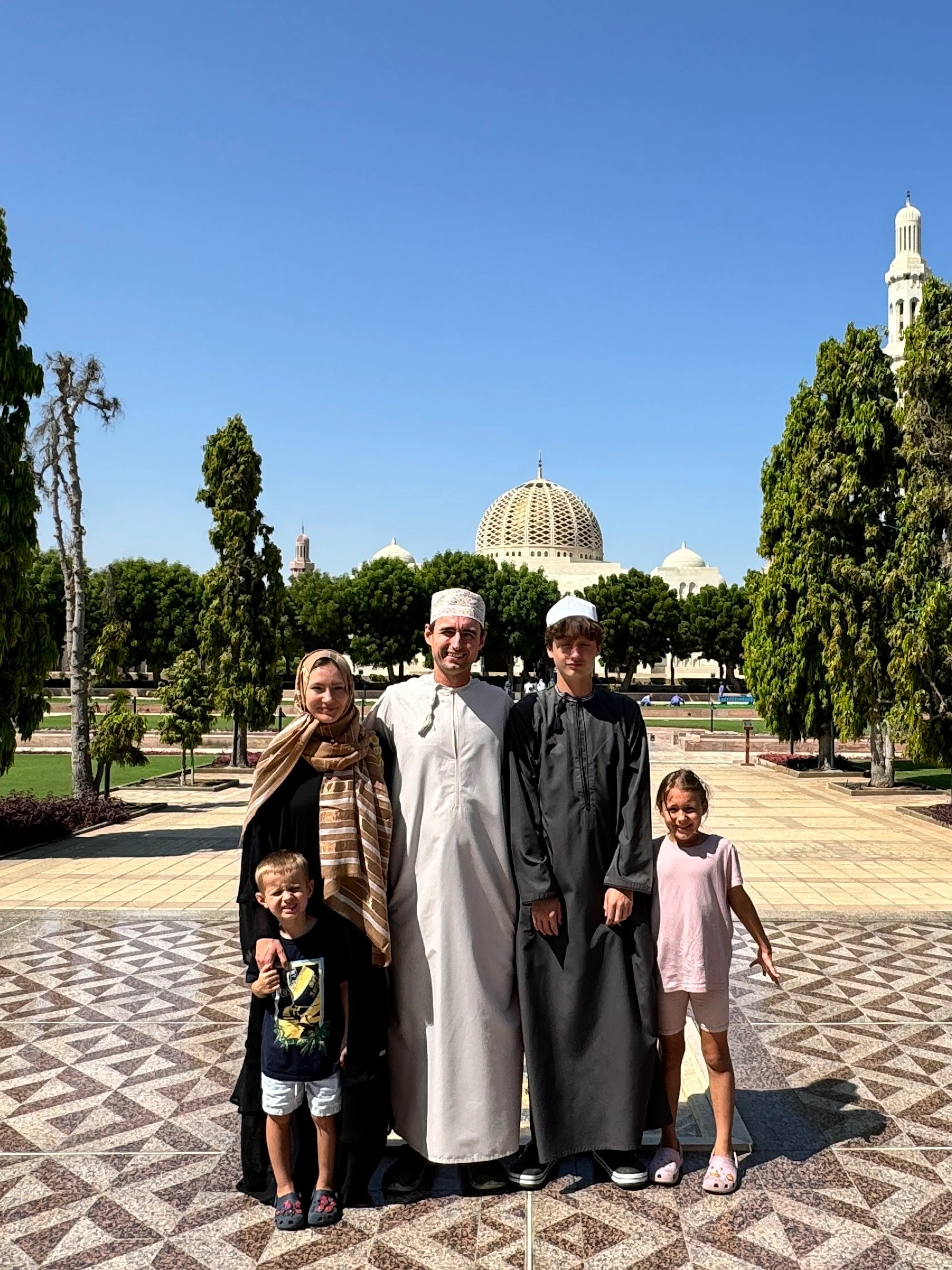 Family standing in front of Sultan Qaboos Grand Mosque, Muscat, Oman, with minarets and gardens.