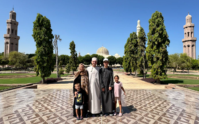 Family standing in front of Sultan Qaboos Grand Mosque, Muscat, Oman, with minarets and gardens.