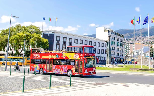 Hop-on hop-off bus in Funchal, Madeira, passing city buildings and flags.