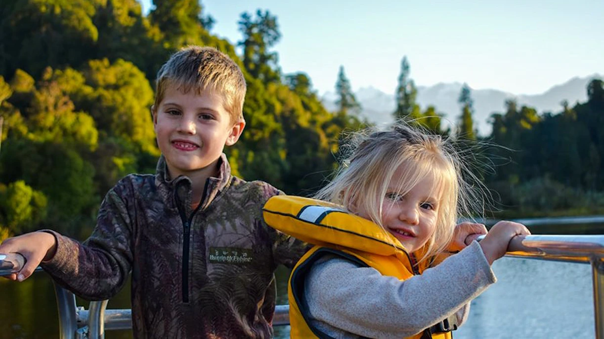 Children enjoying a scenic cruise on Lake Mapourika with forested hills in the background.