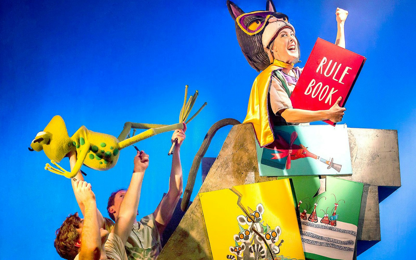 Theater performance of "Oi Frog & Friends!" with puppeteers and character holding a rule book.