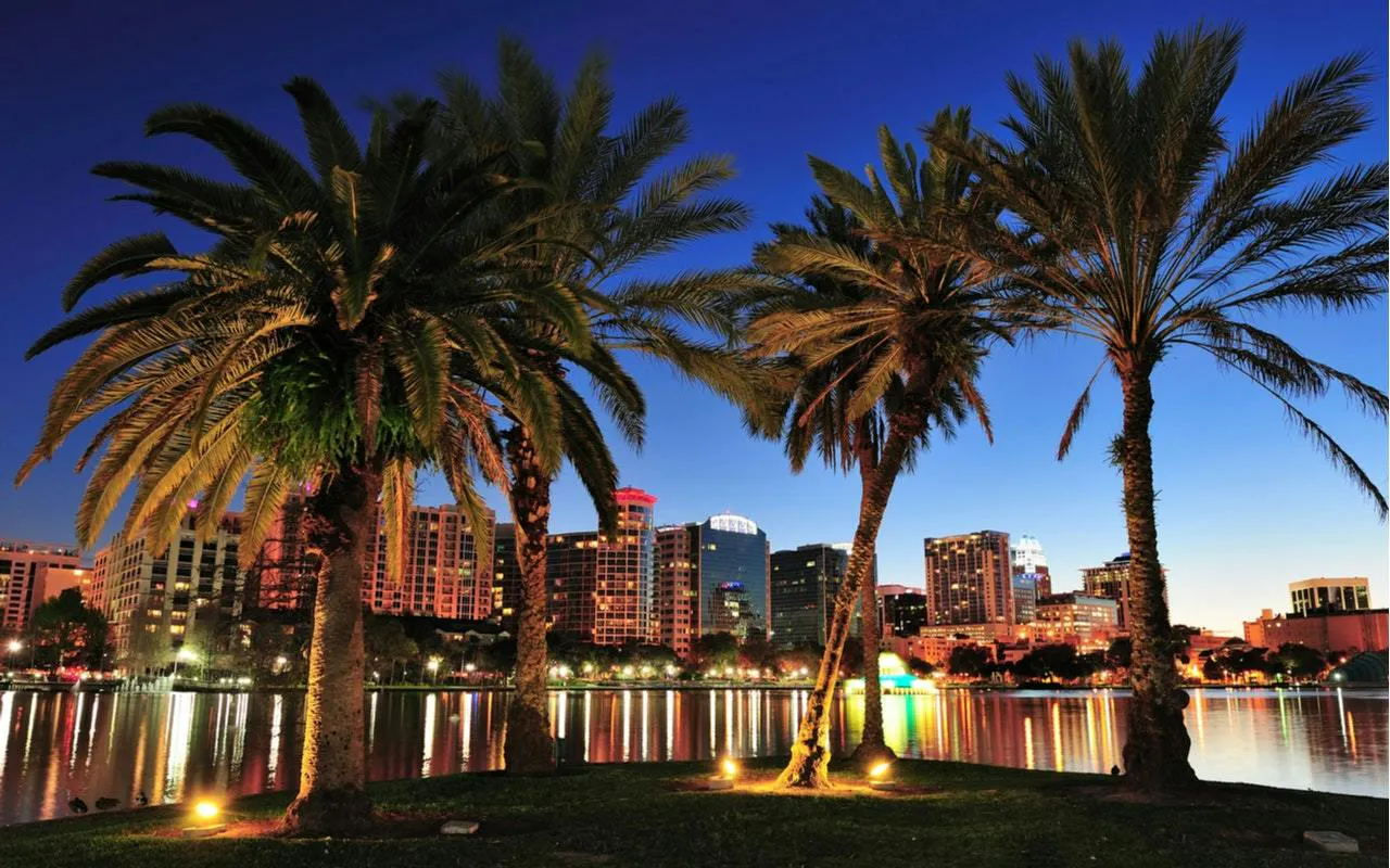 Orlando skyline at dusk with palm trees and city lights reflecting on a lake.