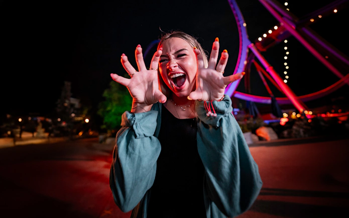Person playfully posing at Six Flags SCarowinds Snicker's Halloween Event with ride in background.