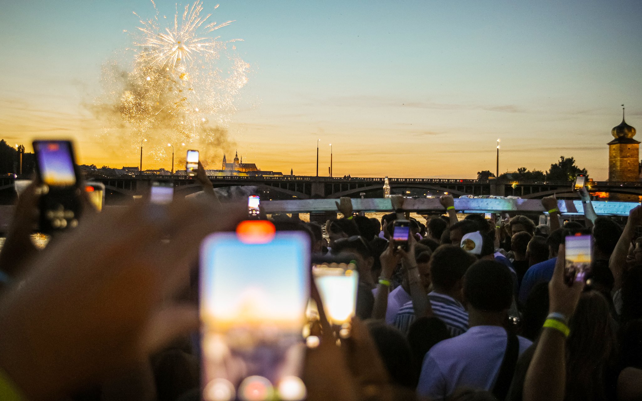 Crowd on boat cruise watching fireworks over river at sunset.