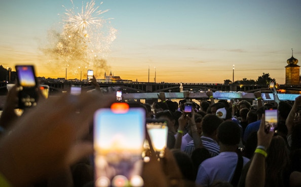Crowd on boat cruise watching fireworks over river at sunset.