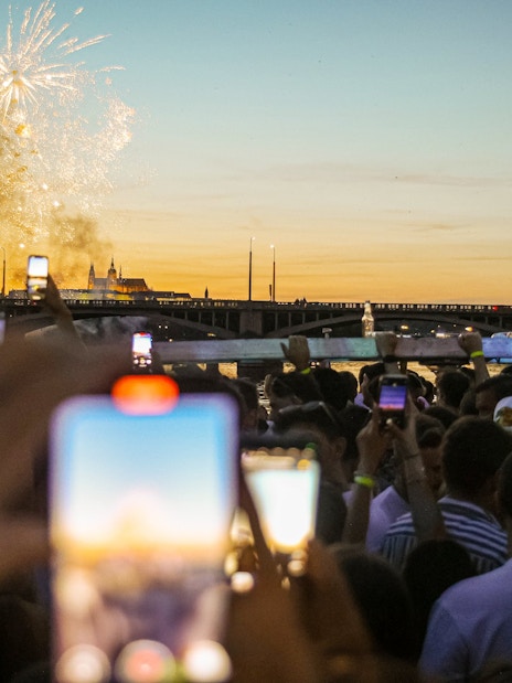 Crowd on boat cruise watching fireworks over river at sunset.