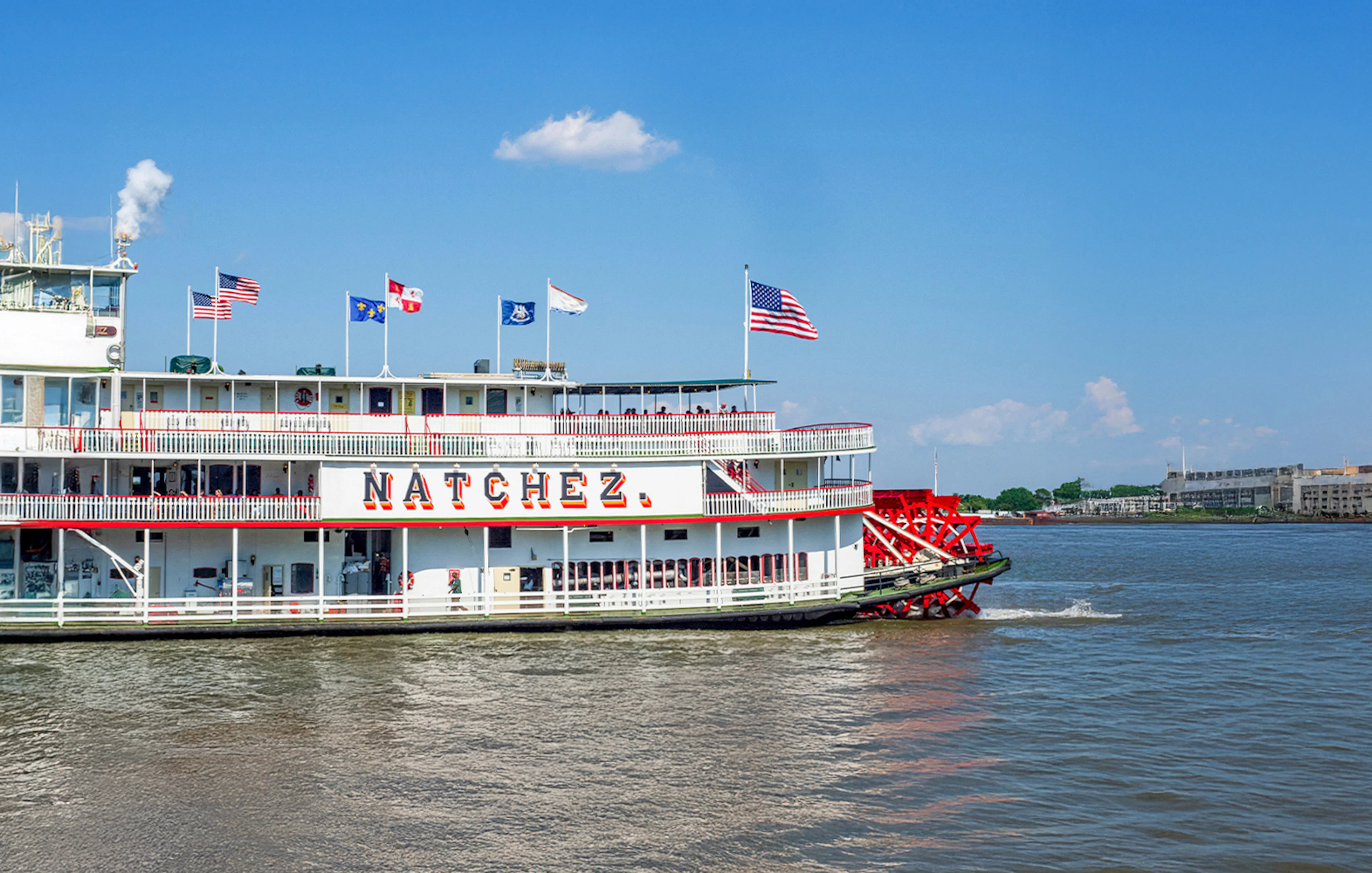 Steamboat Natchez departing from dock on a sunny day with flags flying.