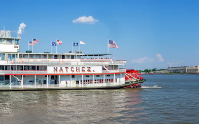 Steamboat Natchez departing from dock on a sunny day with flags flying.