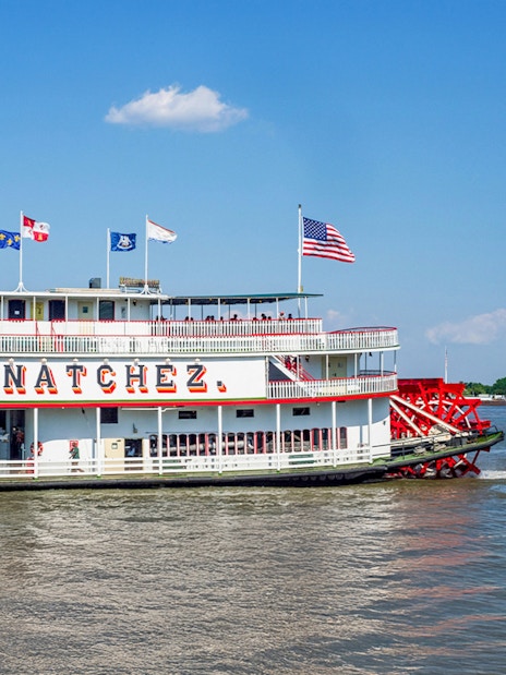 Steamboat Natchez departing from dock on a sunny day with flags flying.