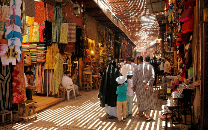 Marrakech market with colorful textiles and people walking through narrow alley.