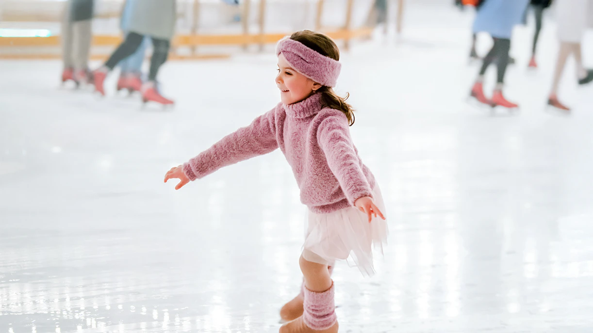 Little girl in pink sweater ice skating indoors.