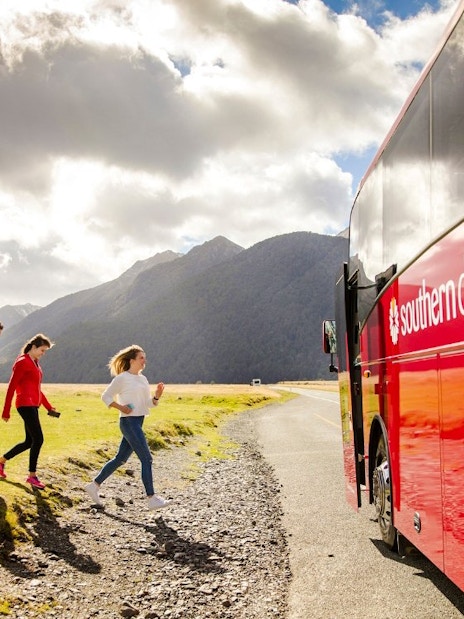 Tourists boarding a red coach to Milford Sound, New Zealand, with mountains in the background.